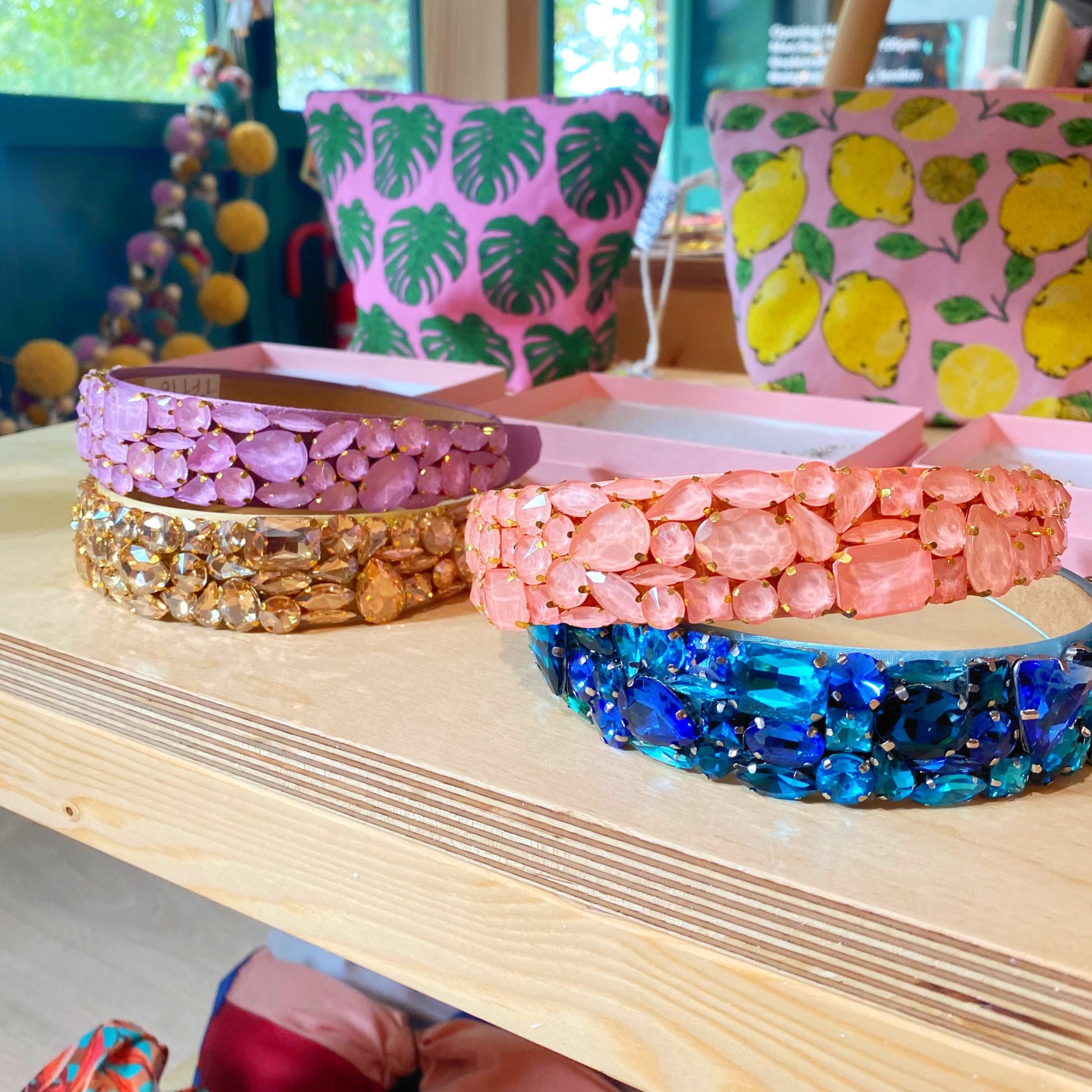 Personalised crystal headbands on display in a store set in the day time. Crystal headbands are on display on a wooden surface with some bags in the background with lemons on. Four crystal headbands on display.